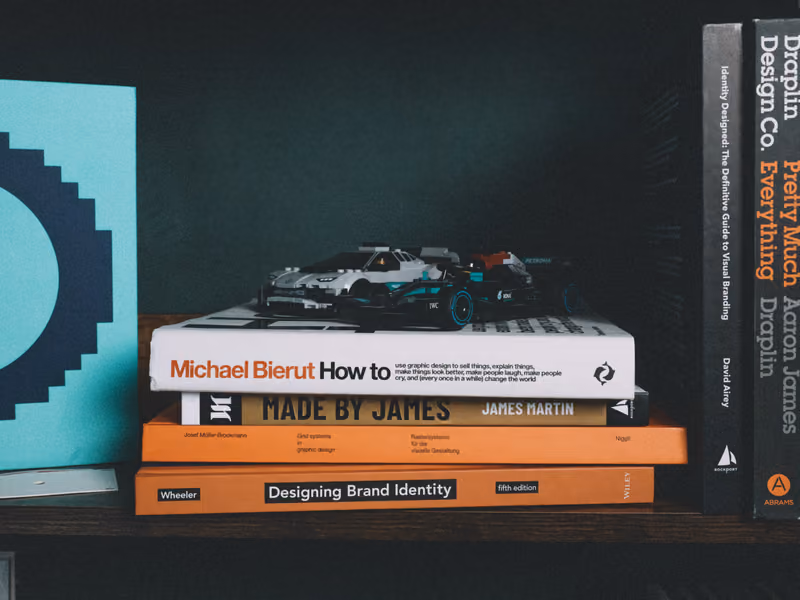 Design books stacked on a shelf with two model racing cars on top.